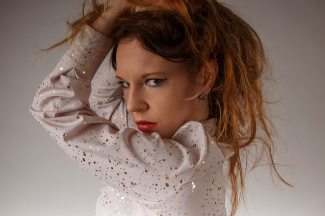 Minimalistyczny portret kobiety o długich, ciemnych rudych włosach, częściowo opadających na twarz. Ma spokojny, poważny wyraz twarzy i subtelny makijaż. Gładkie tło oraz miękkie światło nadają fotografii elegancki, intymny charakter. Kobieca sesja zdjęciowa. Portret Kobiety.