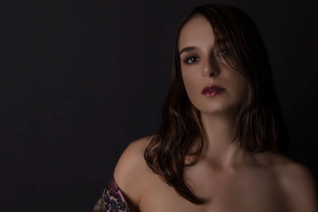 Na zdjęciu widać minimalistyczny portret kobiety o długich, ciemnych włosach, częściowo opadających na twarz. Ma spokojny, poważny wyraz twarzy i subtelny makijaż, a jej ramiona są delikatnie odsłonięte. Ciemne, gładkie tło oraz miękkie światło nadają fotografii elegancki, intymny charakter. Kobieca sesja zdjęciowa. Portret Kobiety.